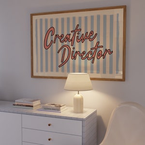May include: A white dresser with three drawers and gold knobs. The dresser has a tiled top with a lamp and books on it. A white chair is in the background. A framed poster with blue and white stripes reads "Creative Director" in pink lettering.