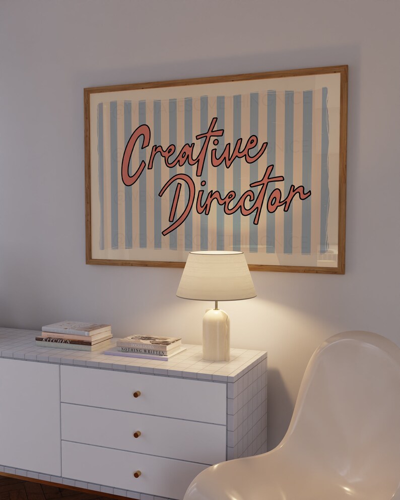 May include: A white dresser with three drawers and gold knobs. The dresser has a tiled top with a lamp and books on it. A white chair is in the background. A framed poster with blue and white stripes reads "Creative Director" in pink lettering.