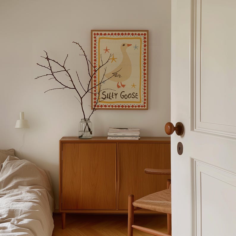Puede incluir: Un armario de madera marr&oacute;n claro con dos puertas, una l&aacute;mpara blanca, un jarr&oacute;n con ramas y una impresi&oacute;n enmarcada de un ganso amarillo con el texto "Silly Goose" sobre un fondo blanco.
