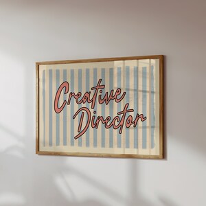 May include: A framed print with a blue and white striped background and the text "Creative Director" in pink cursive lettering.