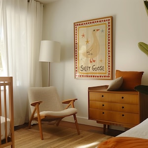Puede incluir: Una silla de guarder&iacute;a blanca con un coj&iacute;n blanco esponjoso se encuentra frente a una c&oacute;moda de madera con tres cajones. Una impresi&oacute;n enmarcada de un ganso de dibujos animados con el texto "Silly Goose" cuelga en la pared sobre la c&oacute;moda.