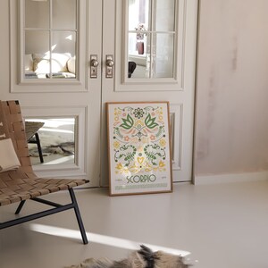 May include: A framed poster with a floral design and the word "SCORPIO" in yellow letters. The poster is on a white floor in a room with white walls and a wooden chair.