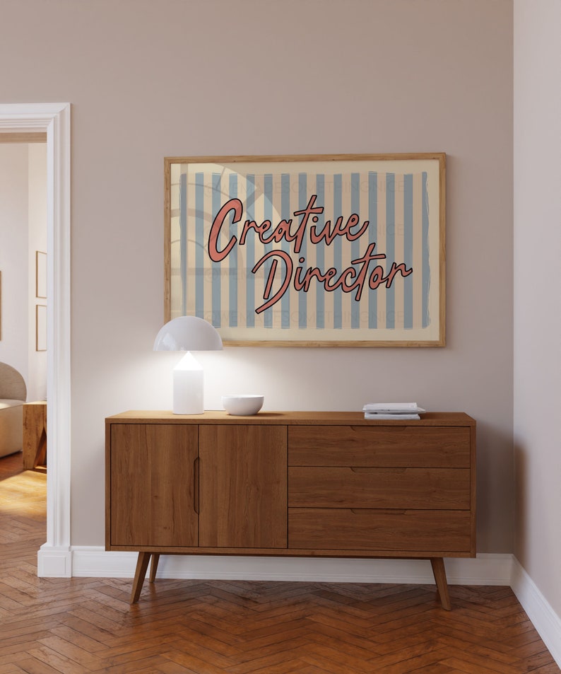 May include: A wooden dresser with three drawers and two doors. The dresser is brown and has a light wood finish. A white lamp and a bowl are on top of the dresser. A framed print with the text "Creative Director" is hanging on the wall above the dresser.
