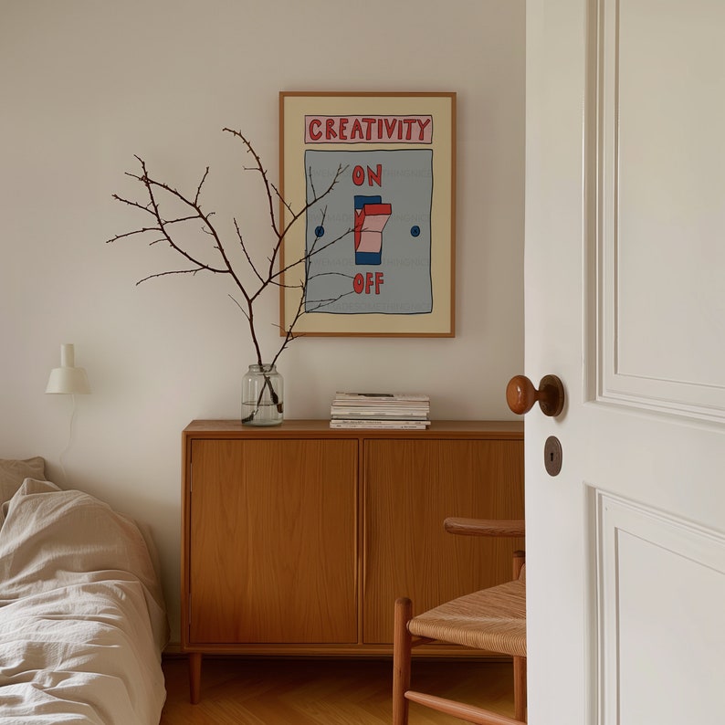 May include: A framed art print with the words "CREATIVITY ON OFF" in red and blue, depicting a light switch. The artwork hangs above a wooden cabinet with a glass vase holding branches. A partially open white door and a woven chair are in the foreground.