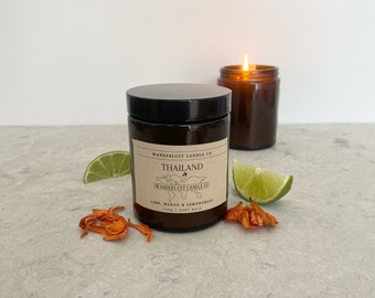 May include: A brown glass candle jar with a black lid. The label reads "Wanderlust Candle Co. Thailand Wanderlust Candle Co. Lime, Mango & Lemongrass". There are lime slices and dried orange flowers on a gray surface.