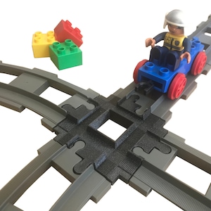 May include: A toy train track set with a blue toy car and a figure of a person wearing a white helmet. The track is made of gray plastic and has a black rubber insert. The car is blue with red wheels and a black steering wheel. The figure is wearing a yellow shirt and a black vest. The figure is holding a black steering wheel.