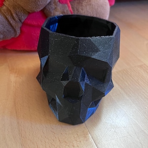 Peut inclure: Un pot en forme de crâne noir et géométrique. Le pot a un design à facettes et un intérieur creux. Le pot en forme de crâne est fait d'un matériau sombre et repose sur une surface en bois. Le pot est un objet décoratif.