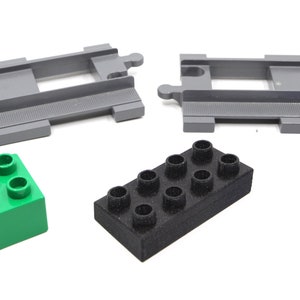 May include: A set of gray plastic train tracks, a green plastic block with four studs, a black plastic block with eight studs, and a blue plastic block with four studs.