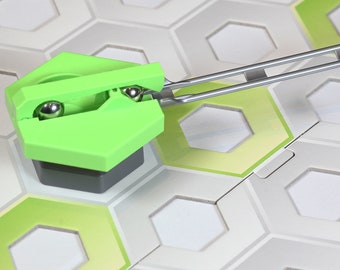 May include: A green and gray game piece with a metal arm attached. The piece has two metal balls inside. The piece is on a white and green hexagonal game board.
