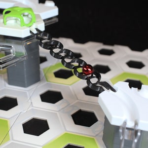Peut inclure: Une chaîne noire avec une bille rouge qui roule dessus. La chaîne est attachée à deux plateformes blanches avec des bases grises. Les plateformes sont sur un plateau hexagonal blanc et vert.