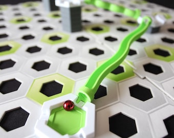 May include: A green plastic track piece with a red marble in a green and white plastic base. The track piece is on a white and green hexagonal game board.