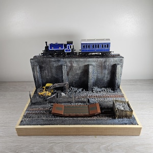 May include: A model train diorama featuring a blue locomotive with the letters CR and a matching blue passenger carriage on an elevated track. Below, a yellow excavator sits near a brown flatbed rail car and a small wooden crate of gravel.