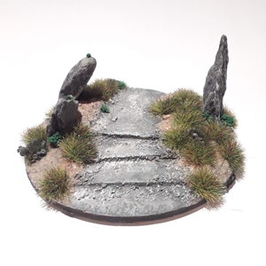 May include: A circular, grey, textured base with a path leading up to two grey rock formations. The base is covered in green and brown grass.