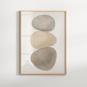 Könnte beinhalten: Gerahmter Kunstdruck mit drei gestapelten, glatten, ovalen Steinen in Beige- und Grautönen. Das Kunstwerk ist in einem hellen Holzrahmen eingefasst, was eine minimalistische und moderne Ästhetik schafft. Die Steine scheinen aquarelliert zu sein.