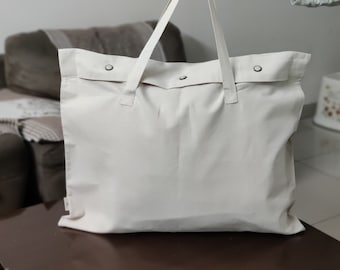 May include: A white canvas tote bag with two silver buttons on the front. The bag has a simple design and is made of a durable material.