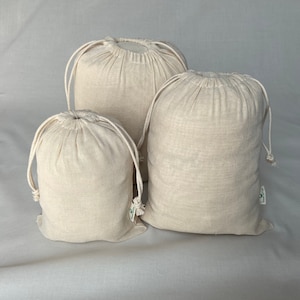 May include: Three off-white cotton drawstring bags of varying sizes. The bags are made of natural cotton fabric and have a simple design.