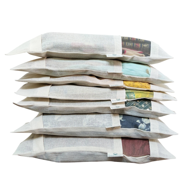 May include: A stack of eight white fabric pillowcases with a small green logo on the bottom right corner. Each pillowcase has a different colorful fabric panel visible through a window in the front. The fabric panels feature various patterns and designs.