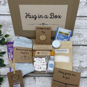 May include: A 'Hug in a Box' gift set in a cardboard box. Contents include a pair of gray socks, a keychain, a tea bag, bath salts, a candle, chocolate, marshmallows, and a small bottle of bubble bath. The box has the text "Hug in a Box".