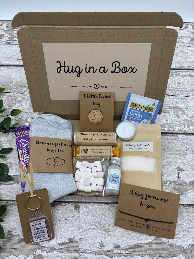 May include: A 'Hug in a Box' gift set in a cardboard box. Contents include a pair of gray socks, a keychain, a tea bag, bath salts, a candle, chocolate, marshmallows, and a small bottle of bubble bath. The box has the text "Hug in a Box".