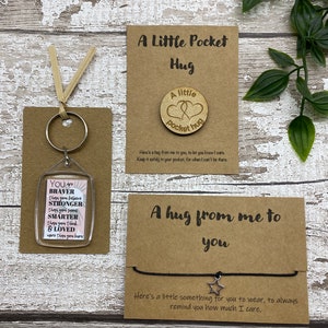 May include: Three small gift items on a wooden surface. Includes a keychain with inspirational text, a wooden pocket hug, and a bracelet with a star charm. The items are on brown cards with text.