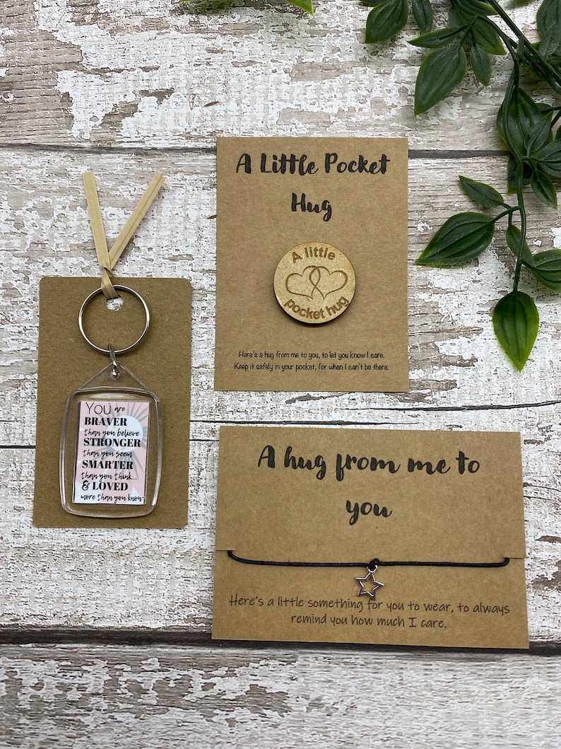 May include: Three small gift items on a wooden surface. Includes a keychain with inspirational text, a wooden pocket hug, and a bracelet with a star charm. The items are on brown cards with text.