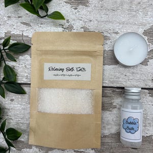 May include: A spa-themed set featuring a kraft paper pouch labeled "Relaxing Bath Salts", a small bottle of "Bubble Bath", and a white tea light candle. The items are arranged on a rustic wooden surface with green foliage.
