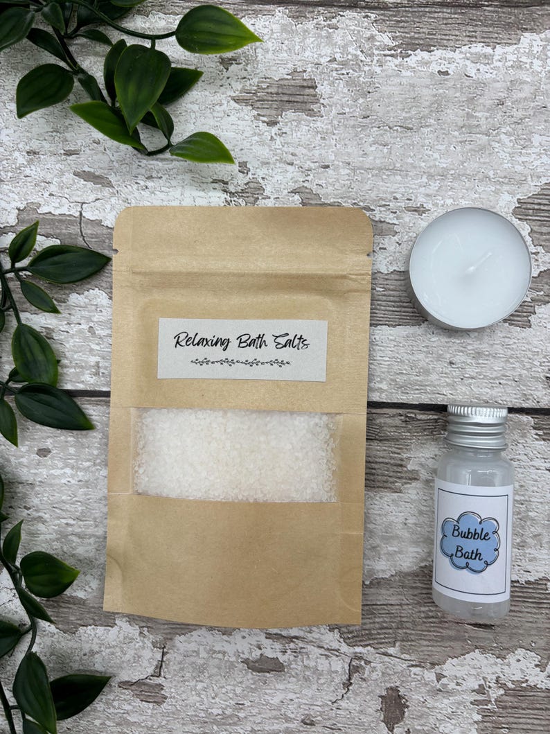 May include: A spa-themed set featuring a kraft paper pouch labeled "Relaxing Bath Salts", a small bottle of "Bubble Bath", and a white tea light candle. The items are arranged on a rustic wooden surface with green foliage.