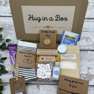May include: A 'Hug in a Box' gift set includes a variety of comfort items. The box contains a pair of striped socks, a chocolate bar, a keychain, bath salts, tea, marshmallows, and a small bottle of bubble bath. The box is brown with the words "Hug in a Box".