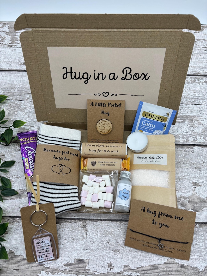 May include: A 'Hug in a Box' gift set includes a variety of comfort items. The box contains a pair of striped socks, a chocolate bar, a keychain, bath salts, tea, marshmallows, and a small bottle of bubble bath. The box is brown with the words "Hug in a Box".