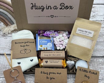 May include: A brown cardboard gift box labeled Hug in a Box containing white socks, hot chocolate, marshmallows, bath salts, a star charm bracelet, a heart-shaped chocolate, a tea bag, and a wooden pocket token, all with printed inspirational messages.
