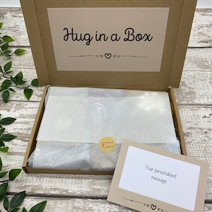 May include: A brown cardboard box labeled "Hug in a Box" with a decorative heart design. Inside, the box contains wrapped contents and a small card that reads "Your personalised message". A sticker reads "handmade with love".