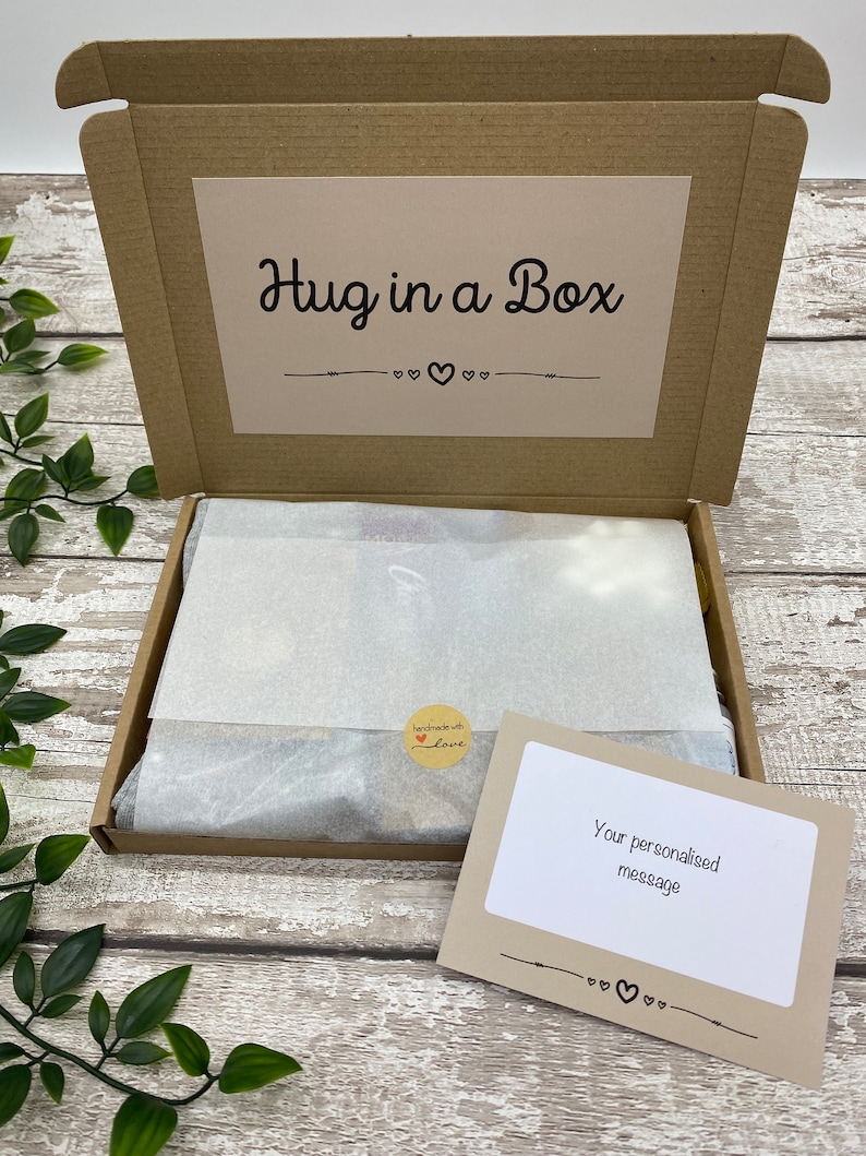 May include: A brown cardboard box labeled "Hug in a Box" with a decorative heart design. Inside, the box contains wrapped contents and a small card that reads "Your personalised message". A sticker reads "handmade with love".