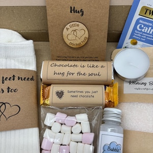 May include: A gift box filled with self-care items. Includes a card that says "A Little Pocket Hug", a wooden token, a chocolate bar, a bag of marshmallows, a bottle of bubble bath, a tea bag, a candle, and a pair of socks.