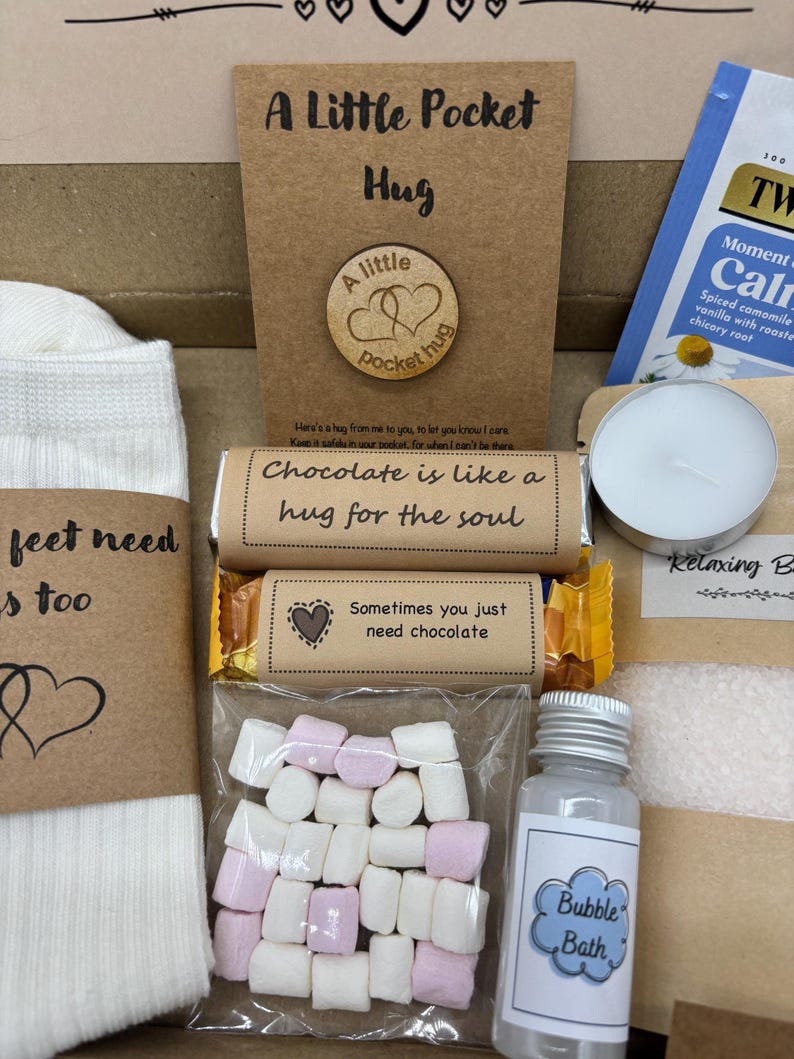 May include: A gift box filled with self-care items. Includes a card that says "A Little Pocket Hug", a wooden token, a chocolate bar, a bag of marshmallows, a bottle of bubble bath, a tea bag, a candle, and a pair of socks.