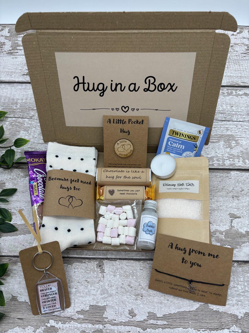 May include: A gift box labeled "Hug in a Box" filled with various items. Includes a chocolate bar, socks, a keychain, bath salts, tea, a small bottle of bubble bath, marshmallows, and a small wooden pin.