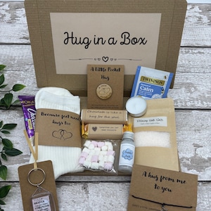 May include: A 'Hug in a Box' gift set includes a variety of comforting items. The box contains a pair of socks, a keychain, a chocolate bar, marshmallows, bath salts, a candle, tea, and a bubble bath. The box is brown with the words "Hug in a Box".