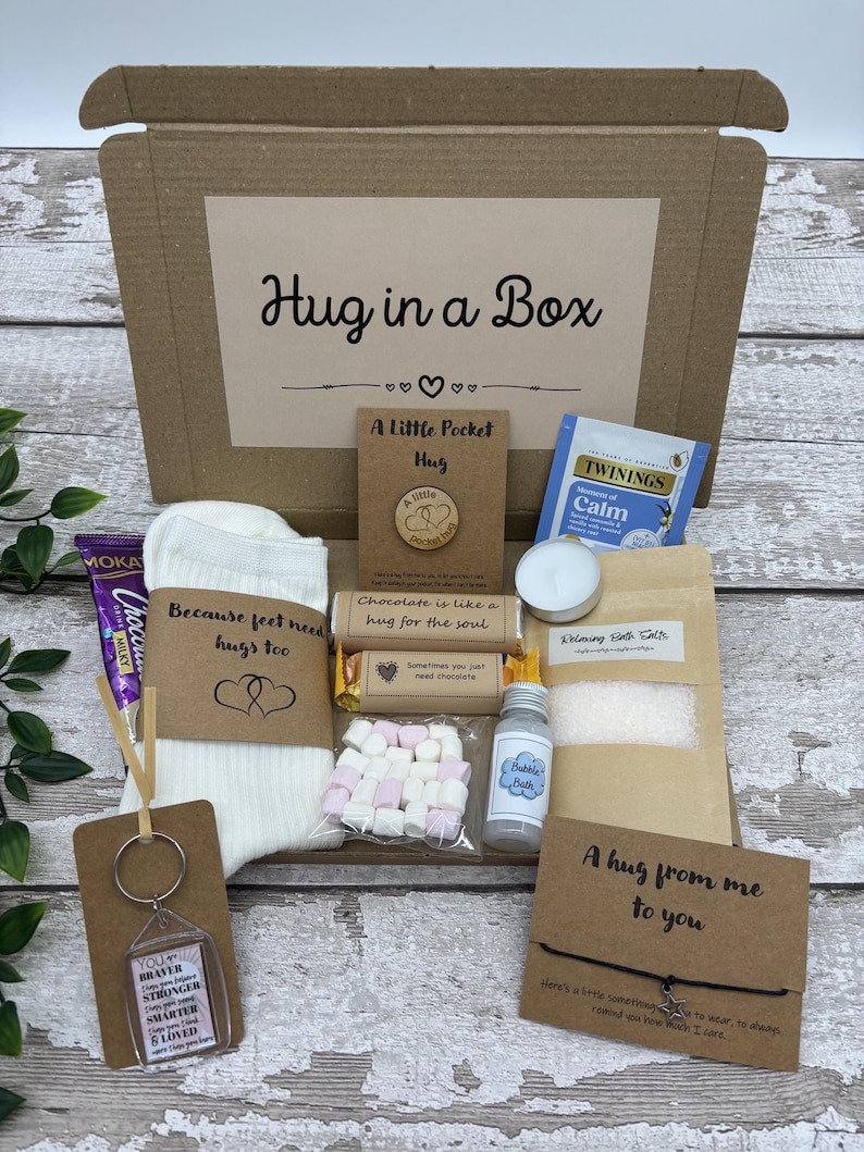 May include: A 'Hug in a Box' gift set includes a variety of comforting items. The box contains a pair of socks, a keychain, a chocolate bar, marshmallows, bath salts, a candle, tea, and a bubble bath. The box is brown with the words "Hug in a Box".