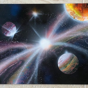 Puede incluir: Una obra de arte realizada con pintura en aerosol que representa una galaxia con una estrella blanca brillante en el centro, rodeada de tres planetas. Los planetas son de color naranja rojizo, morado y verde y marrón. El fondo es negro con rayas de luz blancas y grises.