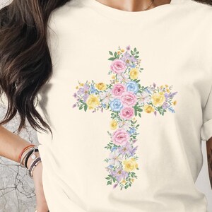 Könnte beinhalten: Cremefarbenes T-Shirt mit floralem Kreuzdesign. Das Kreuz besteht aus pastellfarbenen Blumen, darunter rosa, blaue und gelbe Blüten. Das Shirt hat kurze Ärmel und eine lockere Passform, geeignet für Freizeitkleidung.