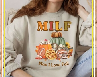 Può includere: Una felpa beige chiaro con la scritta "MILF Man I Love Fall" stampata sul davanti. Il testo è in un font marrone e arancione. Il design presenta un'illustrazione a tema autunnale con zucche, torta e palloni da football.