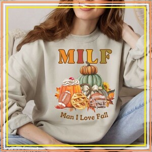 May include: A light beige sweatshirt with the text "MILF Man I Love Fall" printed on the front. The text is in a brown and orange font. The design features a fall-themed illustration with pumpkins, pie, and footballs.