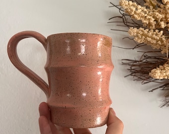 May include: A pink ceramic mug with a speckled glaze and a textured, ribbed design. The mug has a rounded handle.