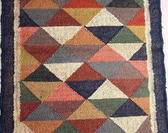 May include: A rectangular jute rug with a geometric pattern of triangles in various colors, including red, green, blue, and beige. The rug has a dark blue border and fringe along the short edges. The rug is approximately 60 inches long and 36 inches wide.