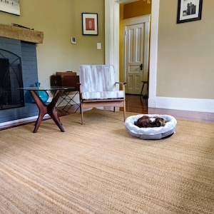 K&ouml;nnte beinhalten: Ein hellbrauner Jute-Teppich mit einem kleinen Hund, der in einem grau-wei&szlig;en Hundebett schl&auml;ft. Der Teppich befindet sich in einem Wohnzimmer mit einem Kamin, einem Holzstuhl und einem Beistelltisch.