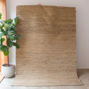 K&ouml;nnte beinhalten: Ein Naturfaser-Teppich mit gewebter Struktur in hellbrauner Farbe. Der Teppich wird an einer wei&szlig;en Wand gezeigt.
