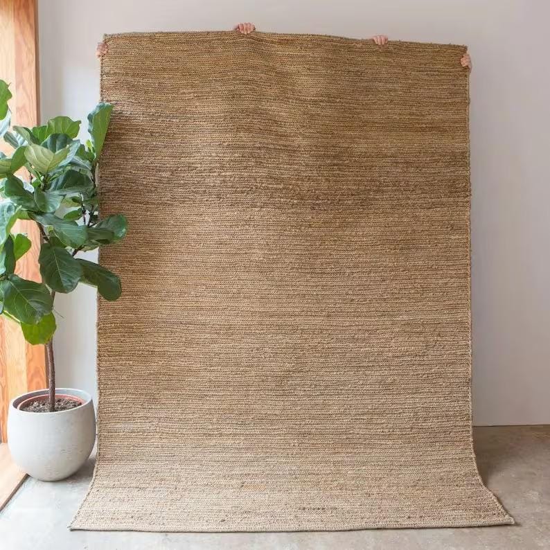 K&ouml;nnte beinhalten: Ein Naturfaser-Teppich mit gewebter Struktur in hellbrauner Farbe. Der Teppich wird an einer wei&szlig;en Wand gezeigt.