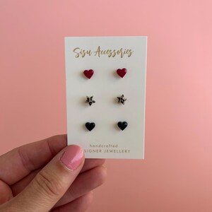 May include: A set of three pairs of stud earrings on a white display card labelled Sisu Accessories. The top pair features red hearts, the middle pair features leopard print stars, and the bottom pair features black hearts. The card is white with gold text.