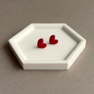 May include: A pair of small, bright red heart-shaped stud earrings. The earrings feature a smooth, solid finish and are positioned on a white, hexagonal-shaped tray with a raised rim, showcasing a simple and clean design for everyday wear.