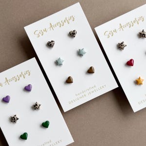 May include: Three white display cards labelled Sisu Accessories featuring various stud earrings in heart and star shapes. The earrings include leopard print, solid purple, green, blue, brown, red, and yellow designs with a glossy finish.