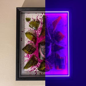 May include: A framed artwork featuring a shattered champagne bottle with green glass shards and pink paint splatters. The right side of the frame is illuminated with a purple neon light.